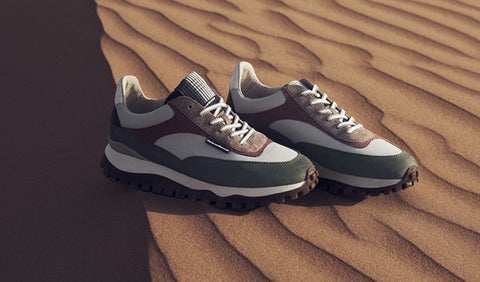 A pair of Floris van Bommel sneakers resting on sand dunes. The shoes feature a colour-blocked design with dark green, white, brown suede, and black soles.