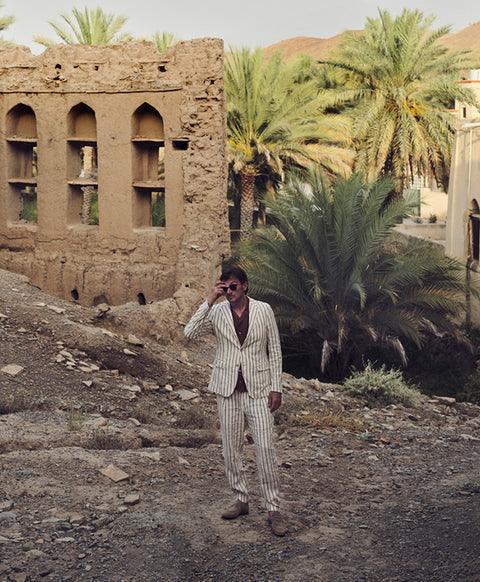 Floris van Bommel wearing a light beige pinstripe suit and sunglasses, stands on a rocky path in front of a desert oasis scene with palm trees and a mud-brick building.