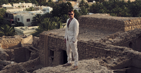 Floris van Bommel wearing a light beige pinstripe suit, brown sunglasses, and brown shoes, stands on a sandy stone structure with desert buildings and palm trees in the background.