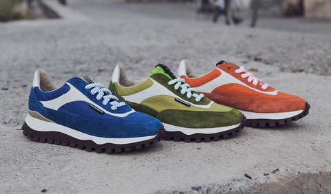Three Floris van Bommel sneakers in blue, olive green, and orange suede and textile blends, arranged side by side on a concrete surface outdoors.