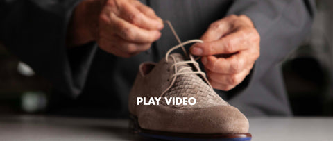 A close-up of a person's hands tying the laces of a taupe suede derby shoe. The person is wearing a dark gray suit jacket, and the background is a dark, blurred indoor setting. Floris van Bommel