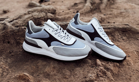 A pair of Floris van Bommel sneakers in light grey, white, and black, styled on a textured brown rocky surface.