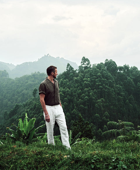 Floris van Bommel stands on a grassy hillside overlooking a dense green forest and misty mountains under a cloudy sky, wearing a dark brown short-sleeve button-up shirt and off-white trousers.