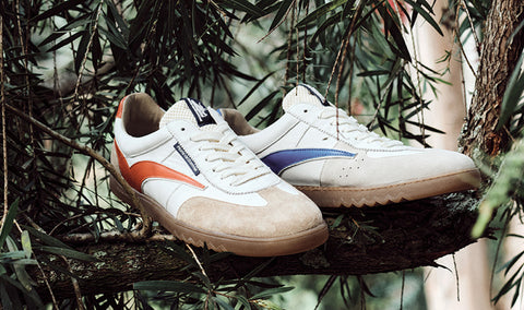 A pair of white Floris van Bommel sneakers with orange and blue side stripes, displayed on a tree branch surrounded by green foliage.