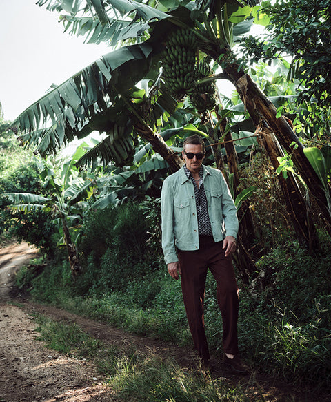 Floris van Bommel wearing a light sage green denim jacket, dark brown trousers, and sunglasses, stands on a dirt path surrounded by lush green banana trees and tropical foliage.