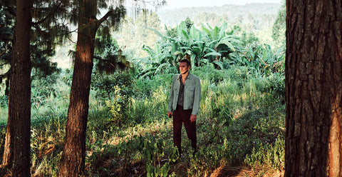 Floris van Bommel, wearing a light green denim jacket, dark brown pants, and a cap stands in a lush green forest clearing framed by tree trunks.