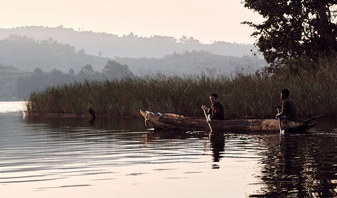 A serene lakeside scene at sunrise or sunset with two people, Floris van Bommel rowing a long wooden boat among tall reeds, with misty hills in the background.
